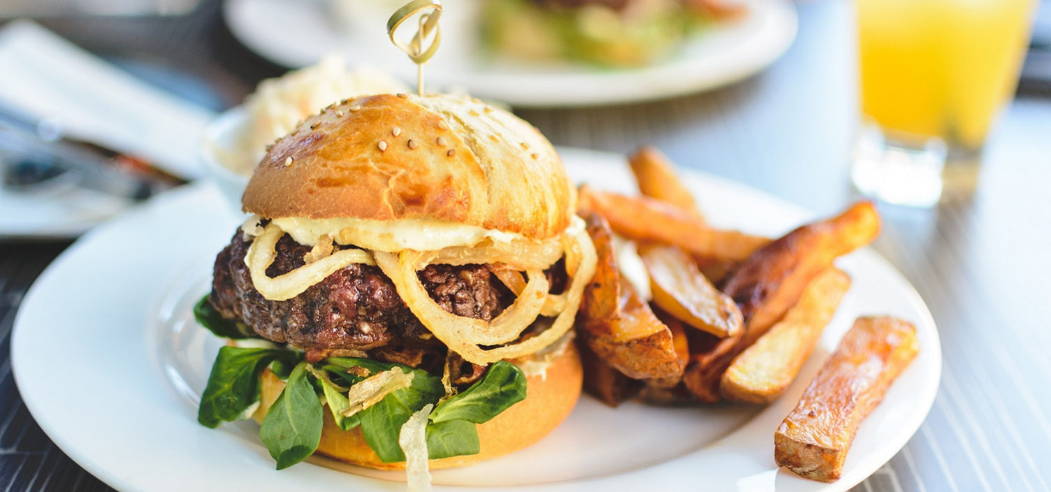 Hamburger mit Steak Fries auf einem weißen Teller. Rechts oben im Hintergrund steht ein Glas mit Orangensaft. Das Geschirr steht auf einem dunkelbraunen Holztisch.