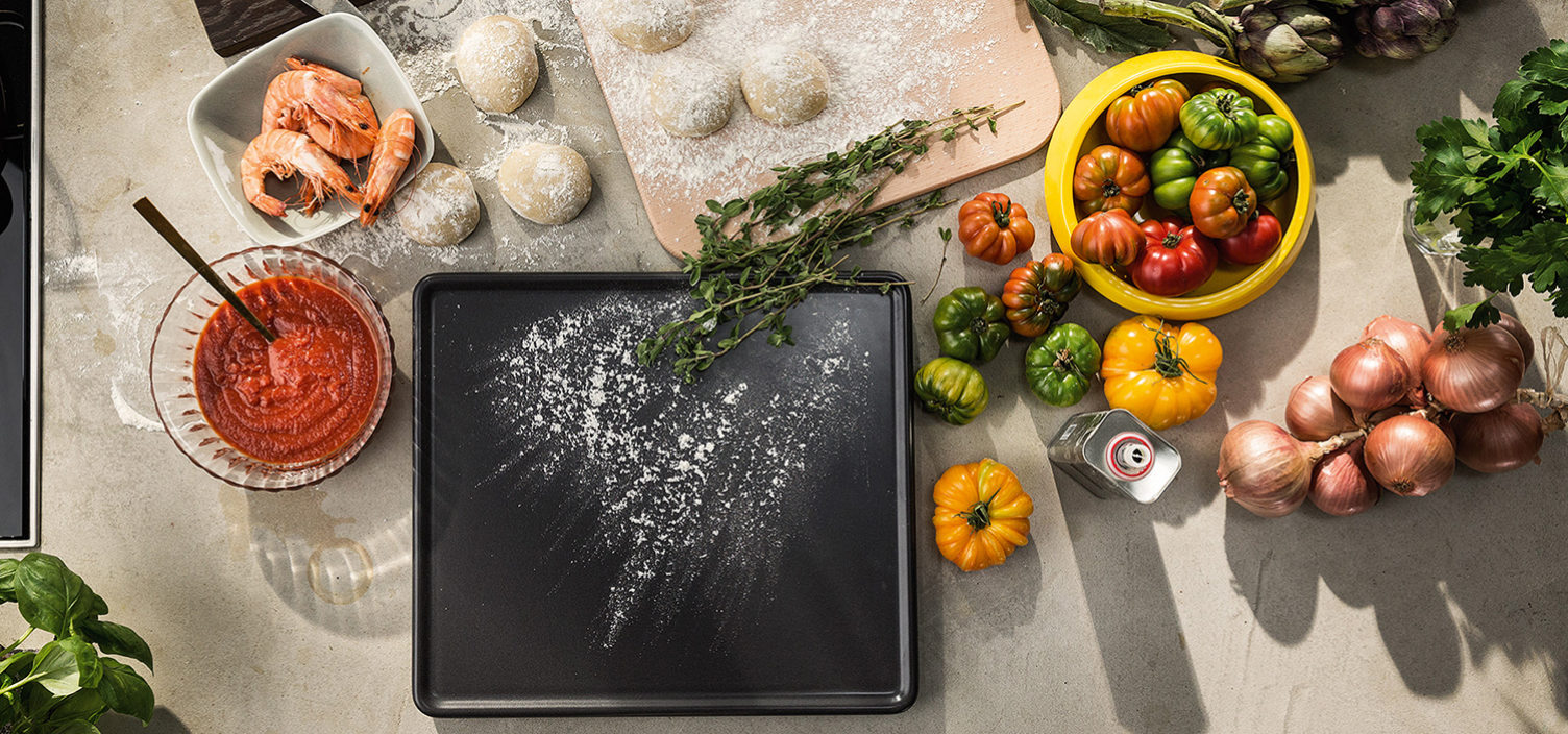 Backstein und Zutaten für die perfekte Pizza auf einem Tisch. Von links nach rechts: Glasschüssel mit Tomatensoße, Garnelen in weißer Schüssel, Pizzateig auf einem Holzbrett, grün, gelb und orange Paradeiser in einer gelben Schüssel und mehrere weiße Zwiebel. Mittig liegt der graue Backstein mit etwas Mehl bestaubt.