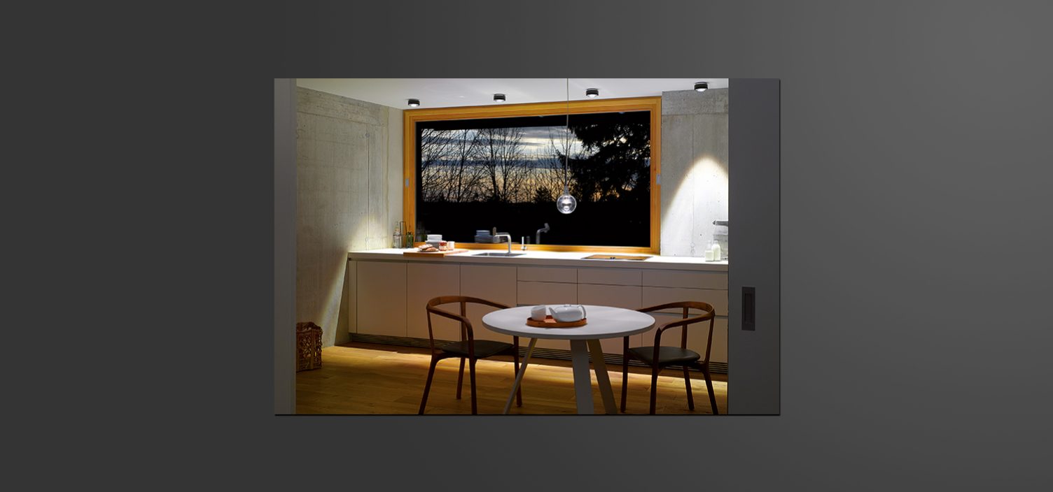 Kleiner runder weißer Tisch mit zwei Sesseln im Vordergrund. Dahinter eine weiße Küchenfront, spärlich beleuchtet mit einem Bild darüber. Auf dem Bild ist ein Wald in der Abenddämmerung zu sehen.