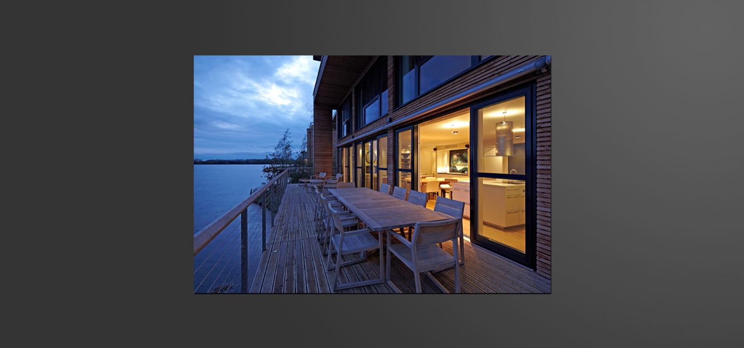 Tafel mit Tischen und Sesseln aus Holz für acht Personen auf einer Außenterrasse neben einem See. Rechts im Bild ist eine Glasschiebetür durch die man ins innere des beleuchtenden Hauses sieht.