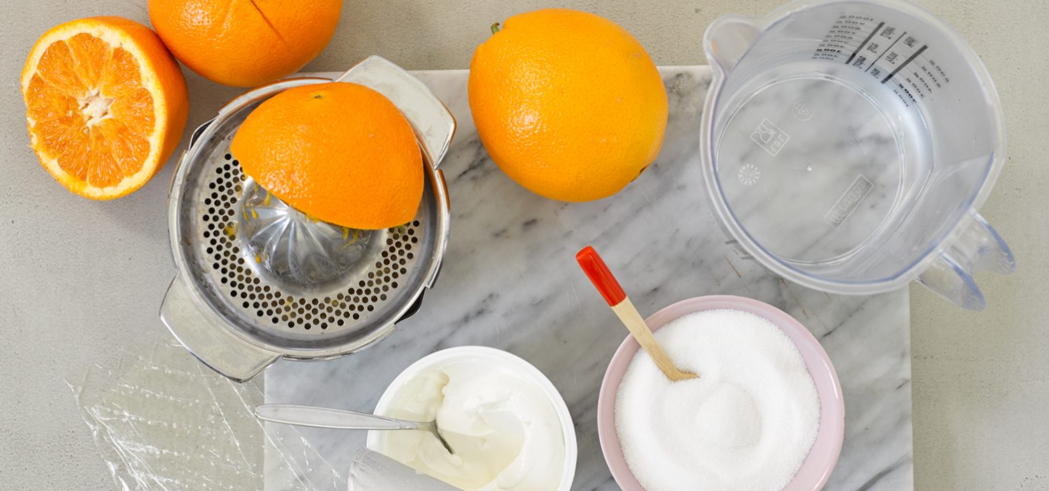 Top-Shot von Zutaten für Orangensorbet: halbierte und ganze Orangen links oben im Bild, darunter zwei Schalen mit Zucker und Schlagobers. Rechts oben im Bild ein Messbecher aus Plastik.