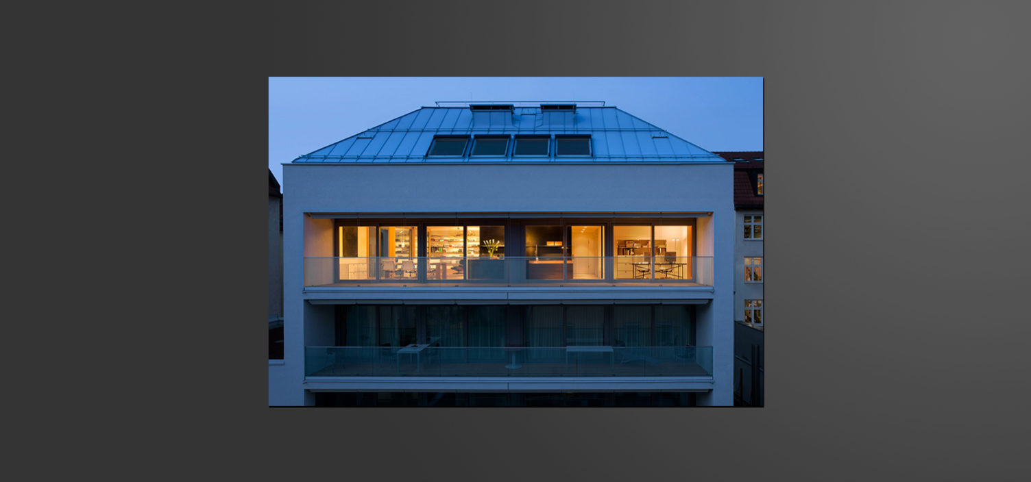 Außenaufnahme von einem Wohnhaus mit Fokus auf das oberste Wohngeschoss mit Außenterasse mit Glasfront. Die oberste Wohnung ist beleuchtet, während in der Wohnung darunter im Bild kein Licht brennt.